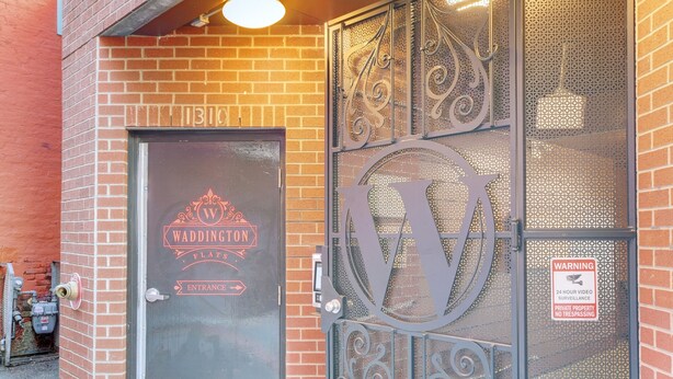 Una reja de hierro forjado con un gran logotipo en forma de 'W', un letrero de advertencia para videovigilancia las 24 horas y una puerta con la inscripción 'WEDDINGTON FLATS ENTRANCE'.