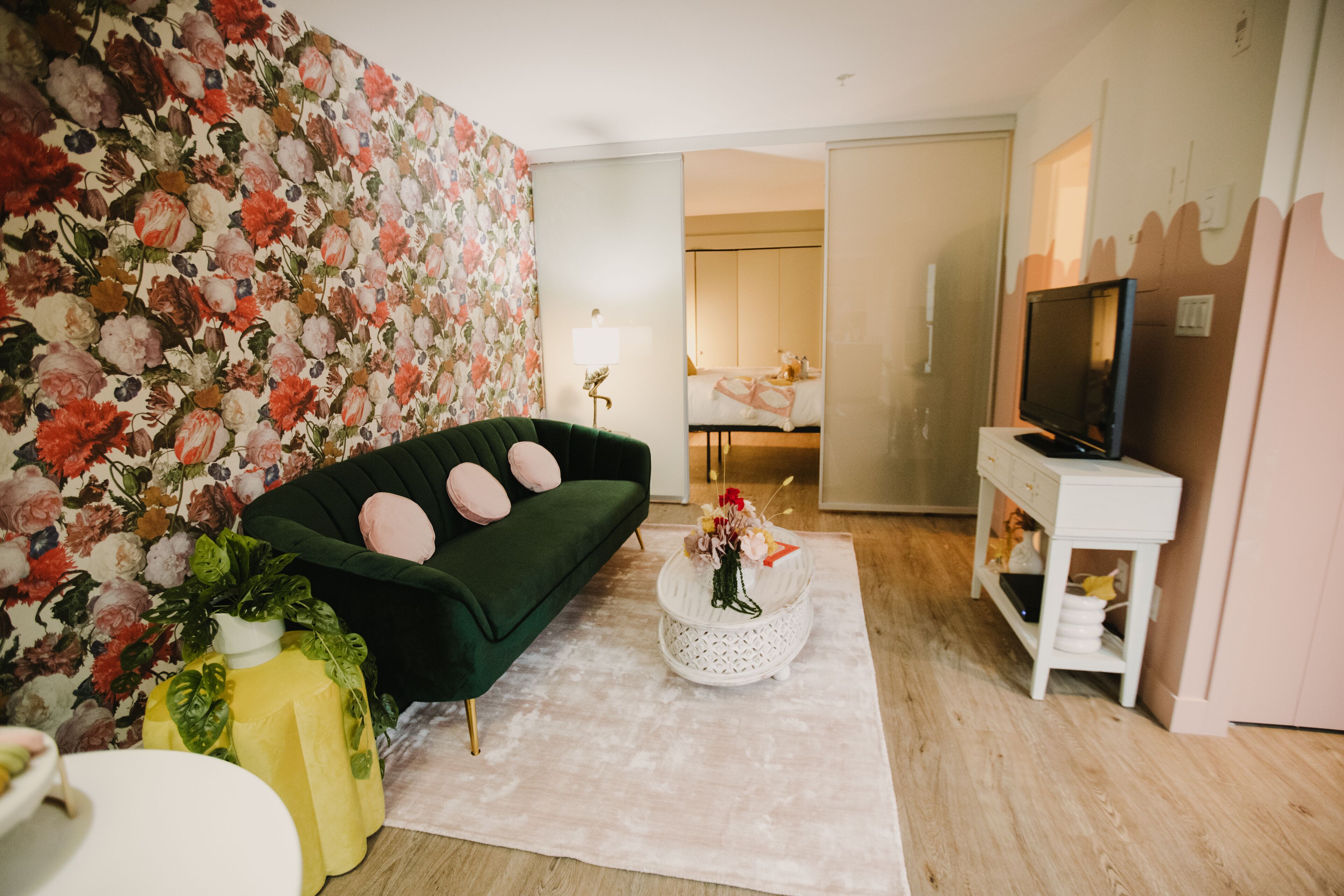 Un salón con papel tapiz floral, un sofá verde con cojines rosados, una mesa de centro blanca con un jarrón, y un televisor sobre un mueble blanco.