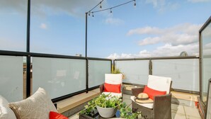 Una terraza en la azotea con barandilla de cristal, mobiliario de mimbre y una planta en maceta.