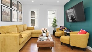 A living room with a yellow sofa, a wooden coffee table, and a TV mounted on a teal wall. - 3BR2Bth TD Garden/BOS/MIT/Harvard/2 Parking Spaces (Somerville)