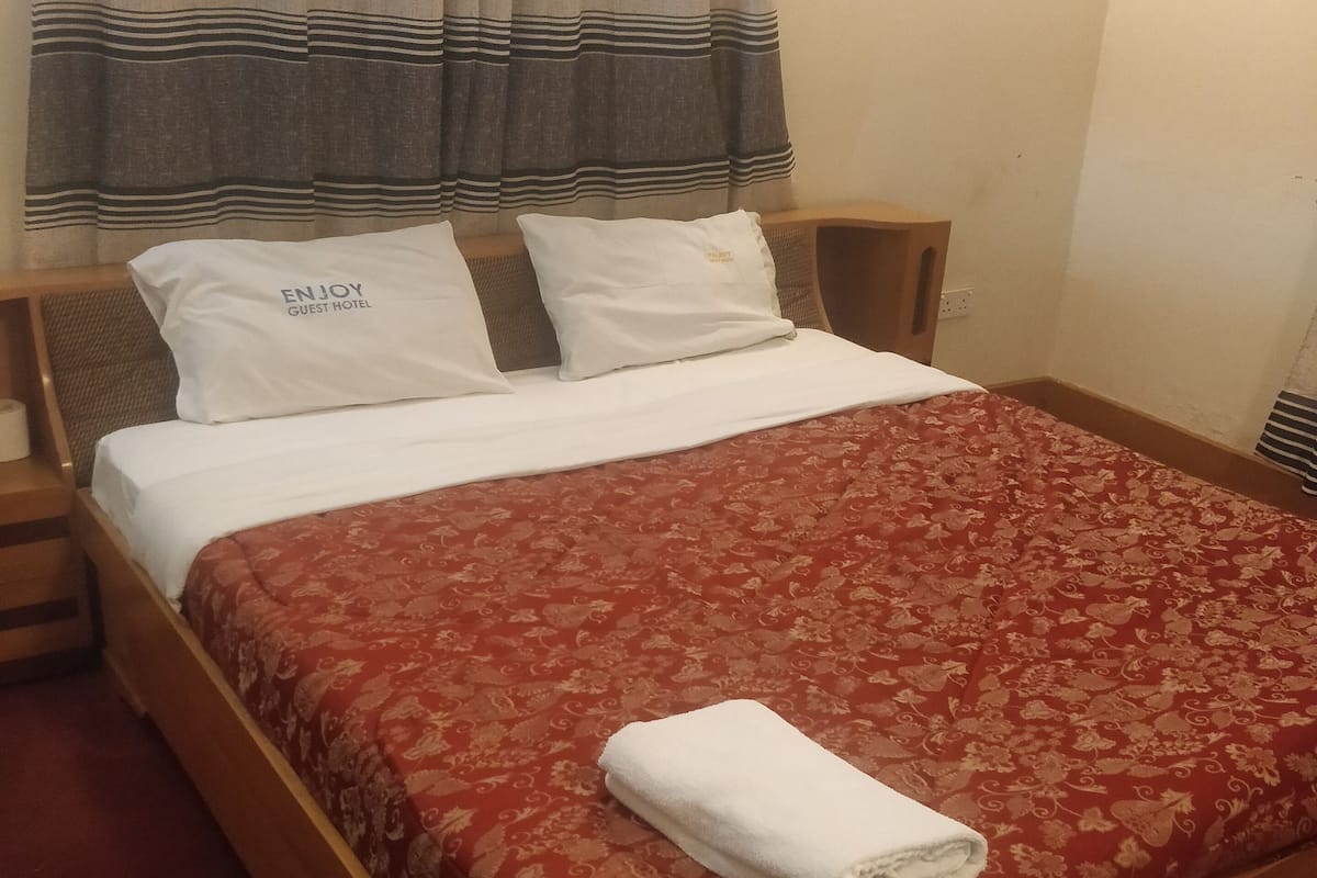 A single bed with a red patterned bedspread, white pillows, and a white towel folded on the bed.