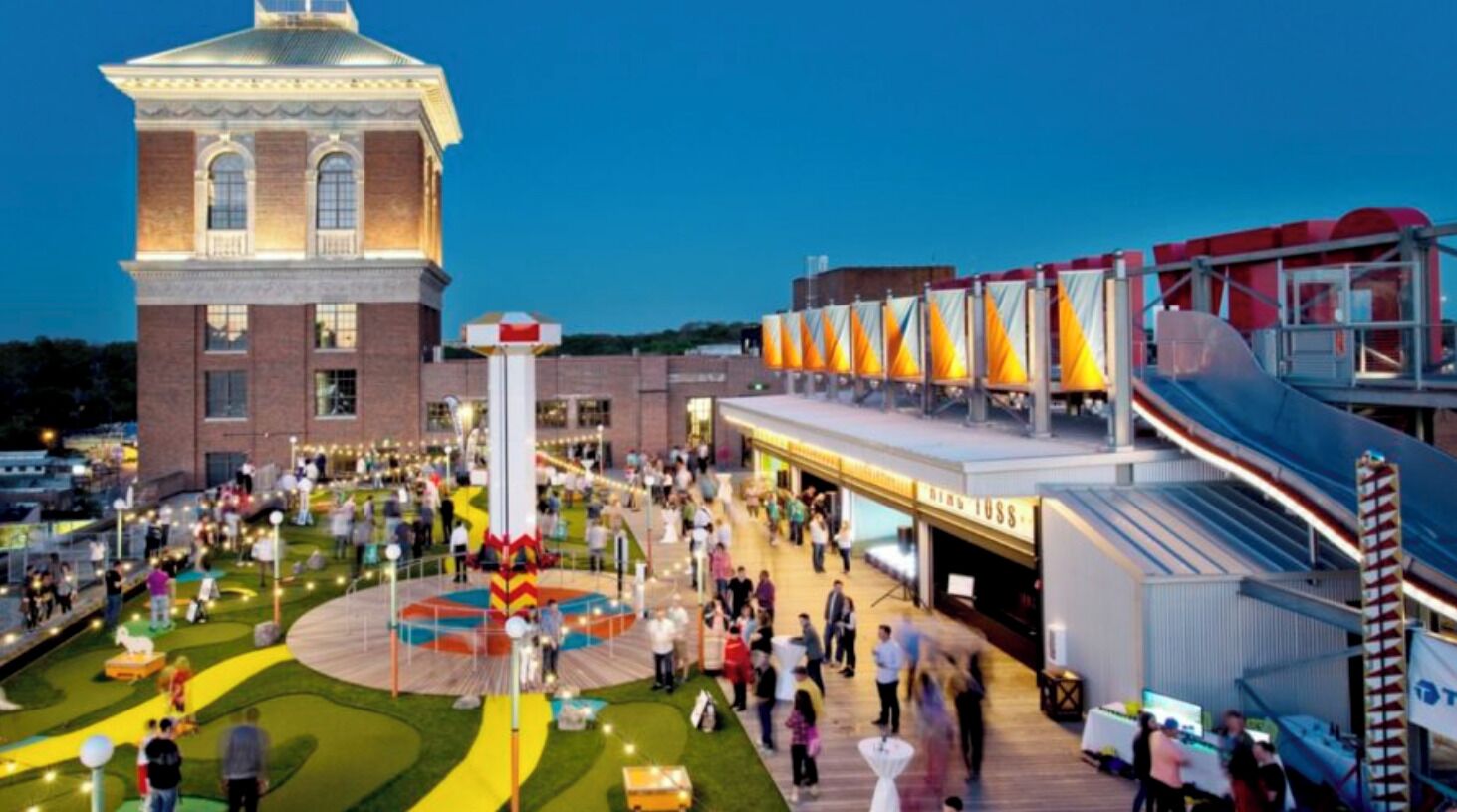 A spacious outdoor area with a central circular garden, a large brick building with a tower, and a modern structure with colorful panels.