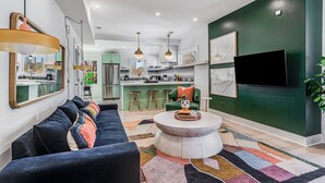 A modern living room with a sofa, coffee table, and a TV. There is a kitchen in the background with stools and a green wall. - Fully Furnished | Fast Wi-Fi | Midterm Stays Welcome (Atlanta)