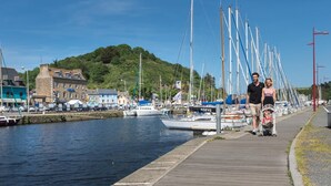 Eine Uferpromenade mit einer Reihe von Booten, die an der Anlegestelle vertäut sind, eine Familie geht vorbei und Gebäude sind im Hintergrund zu sehen.