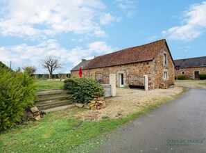 A stone building with a tiled roof, a gravel driveway, and a small garden area with a hedge and steps leading to the entrance. - Le Coin des Hirondelles (HILLION)