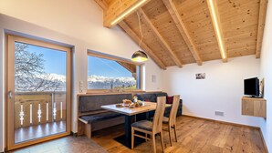 A room with a wooden ceiling, large windows with a mountain view, a dining table with chairs, and a bench.