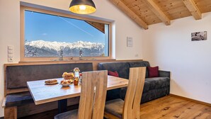 A dining area with a wooden table, chairs, a bench, a large window with a mountain view, and a ceiling with wooden beams.