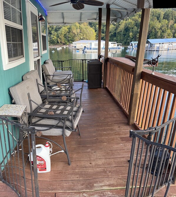 Eine überdachte Terrasse im Freien mit Metallstühlen und einem Tisch, mit Blick auf ein Gewässer mit Booten und Hütten im Hintergrund.