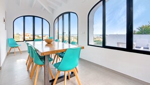 Ein Essbereich mit Holztisch und blaugrünen Stühlen, großen Bogenfenstern und Blick aufs Meer.