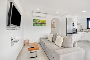 Un saló modern amb un sofà gris, una taula de centre, un televisor penjat i una zona de cuina amb armaris i electrodomèstics blancs.