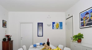 Uma sala de jantar com uma mesa branca, cadeiras, uma garrafa e uma tigela. Há quadros nas paredes e um vaso de planta à direita.