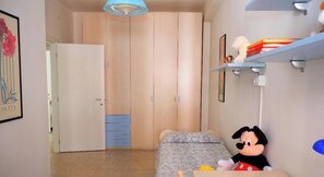 Een slaapkamer met een bed, een Mickey Mouse knuffel, een plank met boeken en een deur die naar een andere kamer leidt.