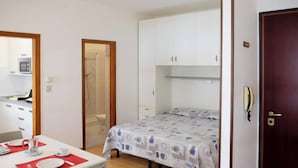 Habitación compacta con cama, una mesita pequeña con un mantel individual rojo, un armario blanco y una puerta con mirilla.