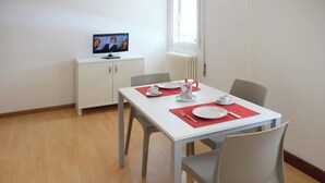Un comedor con una mesa blanca, individuales rojos y un televisor sobre un mueble.