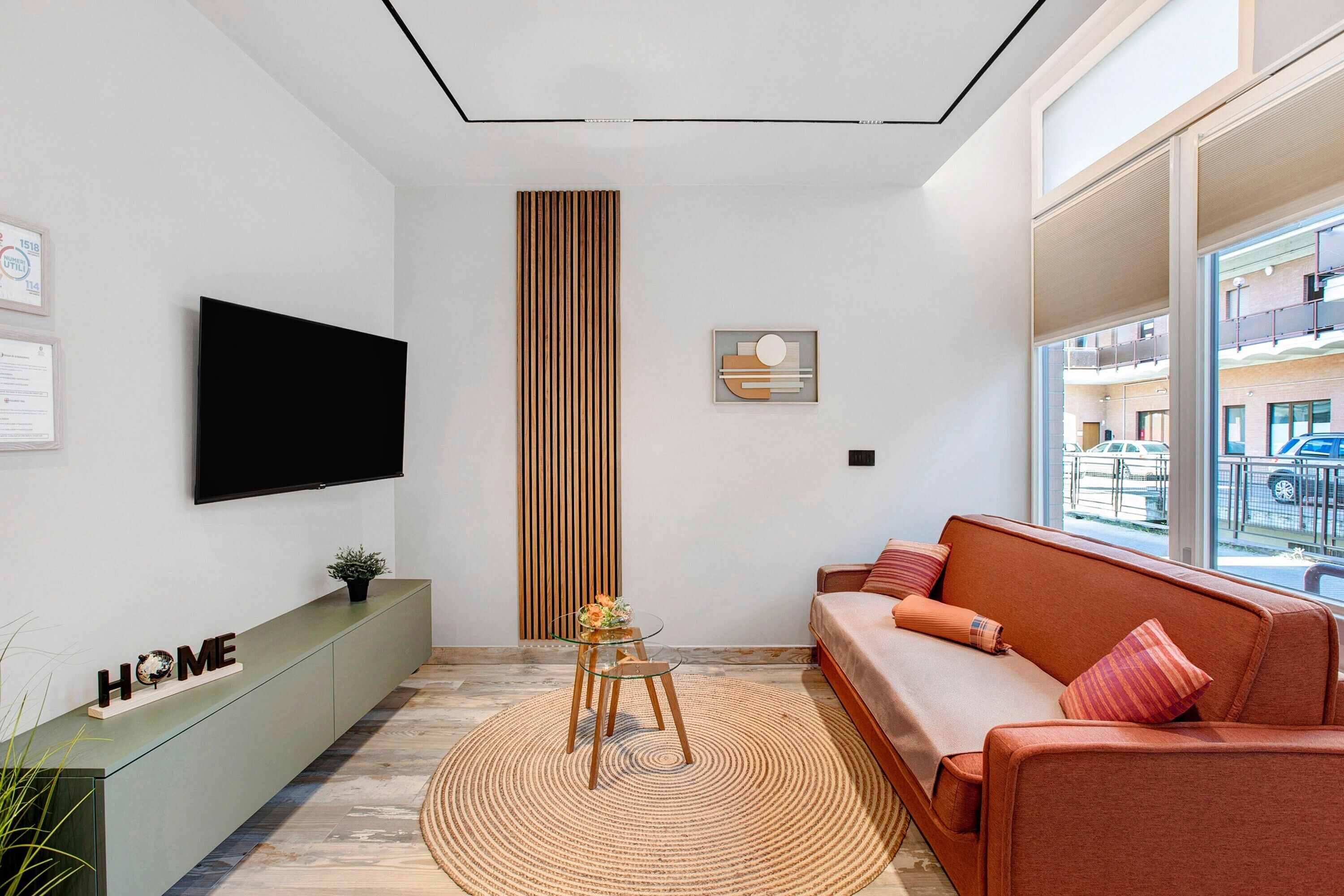 A modern living room with a sofa, a TV, a glass table, a decorative wall panel, and a view through large windows.