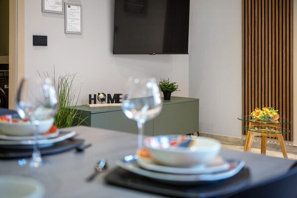 A dining area with a table set for two, a television mounted on the wall, and a small plant on a cabinet.