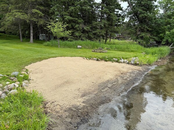 A sandy area with a pond, surrounded by grass and trees.