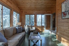 A cozy cabin interior with a sofa, chairs, a table, and large windows offering a view of the forest.