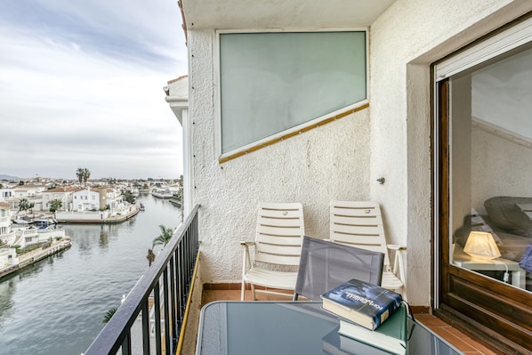 A balcony with two white chairs, a glass table, and a view of a canal with buildings.