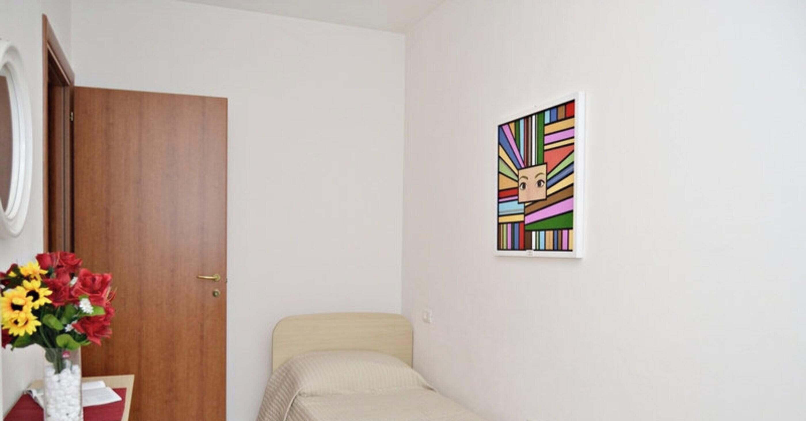 A room with a wooden door, a bed with a white headboard, a wall-mounted picture, and a vase with flowers on a table.