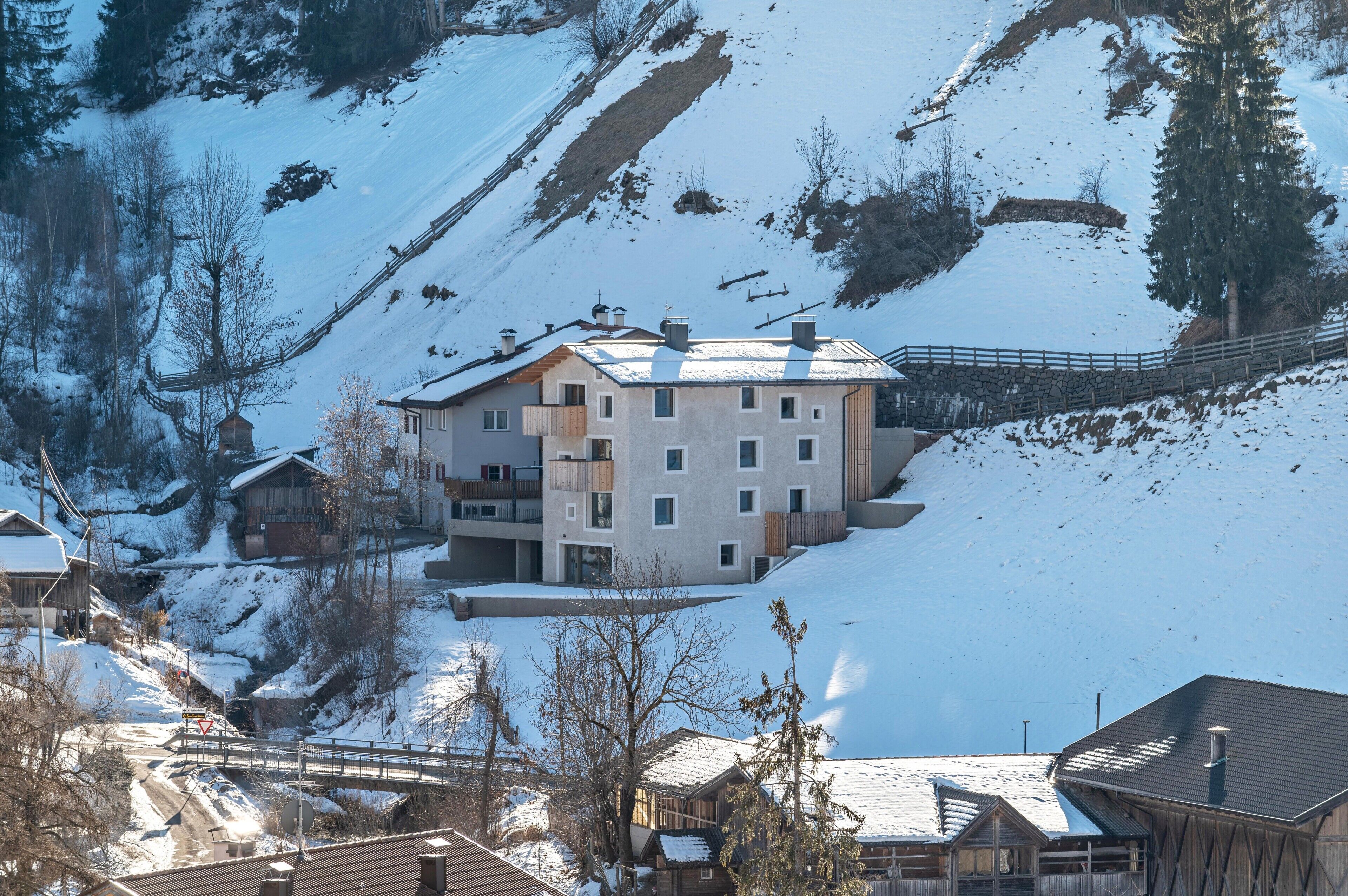 Un edificio de varios pisos con tejado nevado, ubicado en una ladera con cerca de madera y un pequeño puente en primer plano.