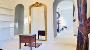 A spacious hallway with white walls, a large ornate mirror, a small wooden chair, and a patterned rug.