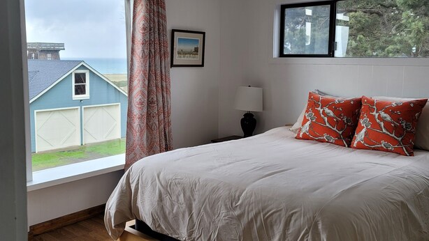 A bedroom with a large bed, a window with a view of a house and the sea, a framed picture on the wall, and a nightstand with a lamp. - Mendocino Ocean View Apartment #6 (Mendocino)