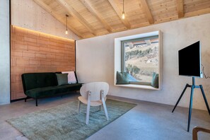 Une pièce avec un plafond en bois, un canapé vert, une chaise blanche, une grande fenêtre donnant sur un paysage enneigé et un téléviseur posé sur un trépied.