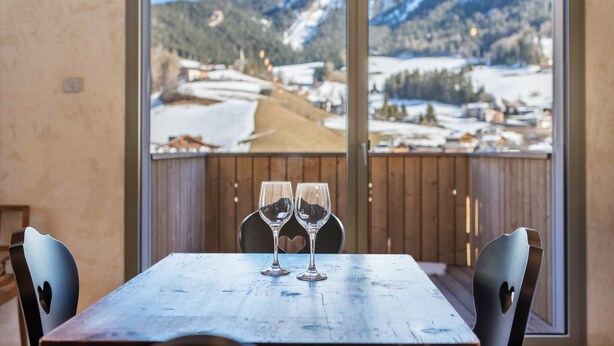 A table with two empty wine glasses, a wooden chair with a heart cutout, and a view of a snowy mountain landscape through a large window. - Apartment 'Lena Dolomites Luxury Suite' with Mountain View, Private Terrace and Sauna (Villnöß)