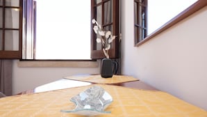 Una habitación con una mesa de color amarillo con estampado, un jarrón con ramas secas y una ventana con marcos de madera.