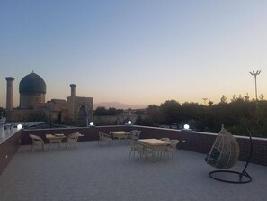 A rooftop terrace with tables and chairs, a hanging chair, and a view of a mosque with a dome and minarets. - Hotel Grand Marakand (Samarkand)