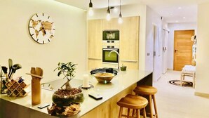 A modern kitchen with a central island, wooden bar stools, a world map clock, and a potted plant.
