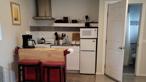 Une cuisine compacte équipée d’une cuisinière, d’un four à micro-ondes et d’un réfrigérateur. On y trouve deux tabourets en bois et une petite table. Une porte donne accès à la salle de bain.