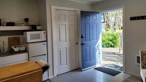 Une cuisine équipée d’un four à micro-ondes, d’un réfrigérateur et d’une planche à découper en bois. Une porte bleue donne accès à un jardin.