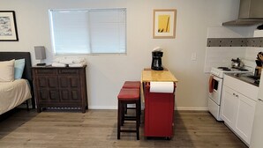 Un espace de vie compact comprenant un coin cuisine, un espace repas avec une banquette, un lit et une fenêtre avec des stores.