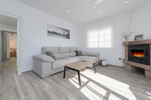 A modern living room with a grey sofa, a wooden coffee table, a fireplace, and a large window. - Lovely vacation home Casa jabe (Cómpeta)