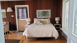 A bedroom with a wooden floor, a bed with white bedding, a desk with a chair, and a window with blinds. - Mendocino 2nd Floor Studio #7 in the Village, full kitchen (Mendocino)