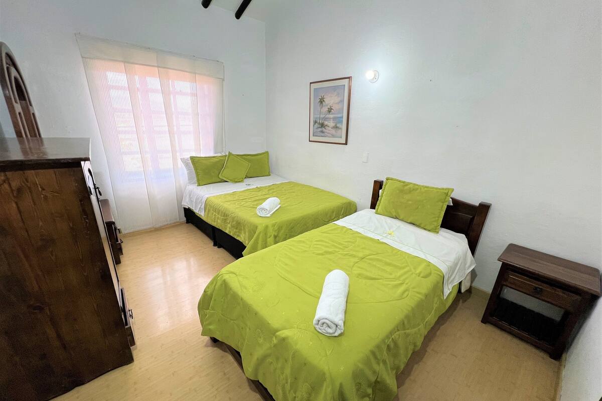 Een kamer met twee bedden, een houten dressoir, een klein nachtkastje en een raam met jaloezieën.