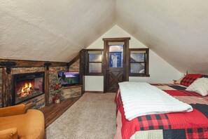 A cozy attic bedroom with a fireplace, a bed with a red and black checkered bedspread, and a television displaying a scenic landscape. - Tahoe Pines Cabin (Homewood)