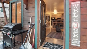 Entrada de uma cabana com churrasqueira e pás na parte externa, sala de estar interna e um letreiro com a inscrição 'BEM-VINDO à cabana'.