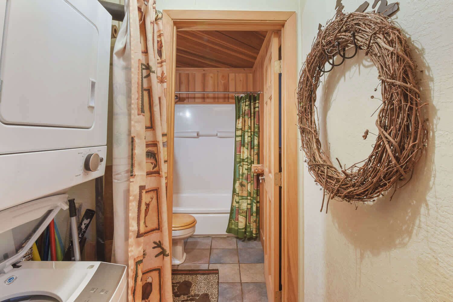 Een wasruimte met een wasmachine, een houten deur naar een badkamer met toilet en douche, en een krans aan de muur.