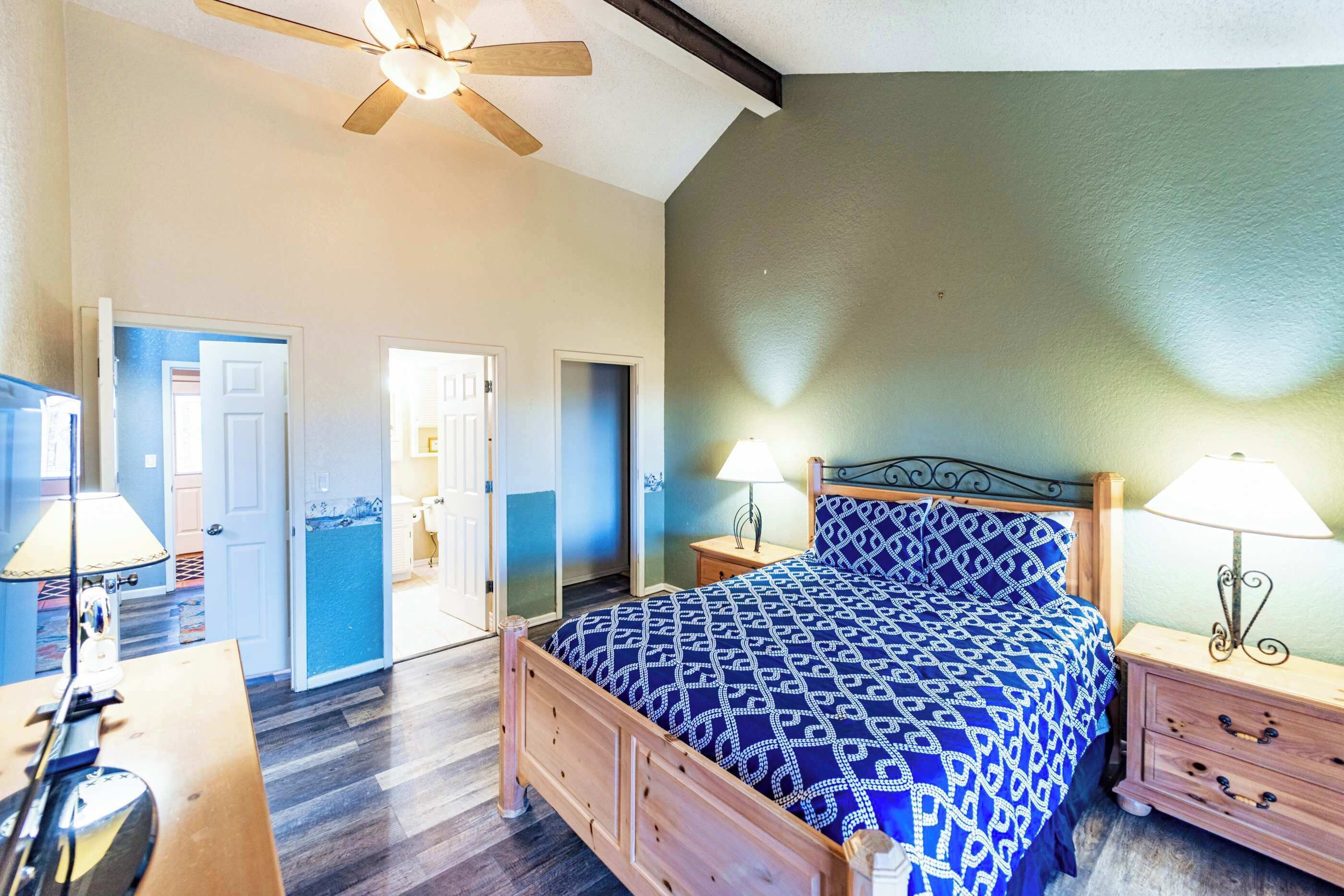 A bedroom with a wooden bed, two bedside tables with lamps, a ceiling fan, and a door leading to another room.