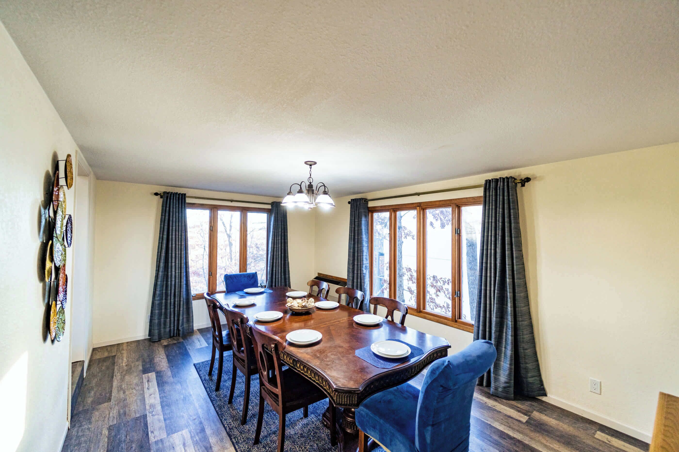 Een eetkamer met een houten tafel voor zes personen, omringd door stoelen, grote ramen met gordijnen en een kroonluchter.