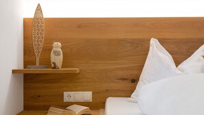 A neatly made bed with white bedding, a wooden nightstand with a book and a decorative item, and a wall-mounted shelf with a wooden ornament and a small figurine.