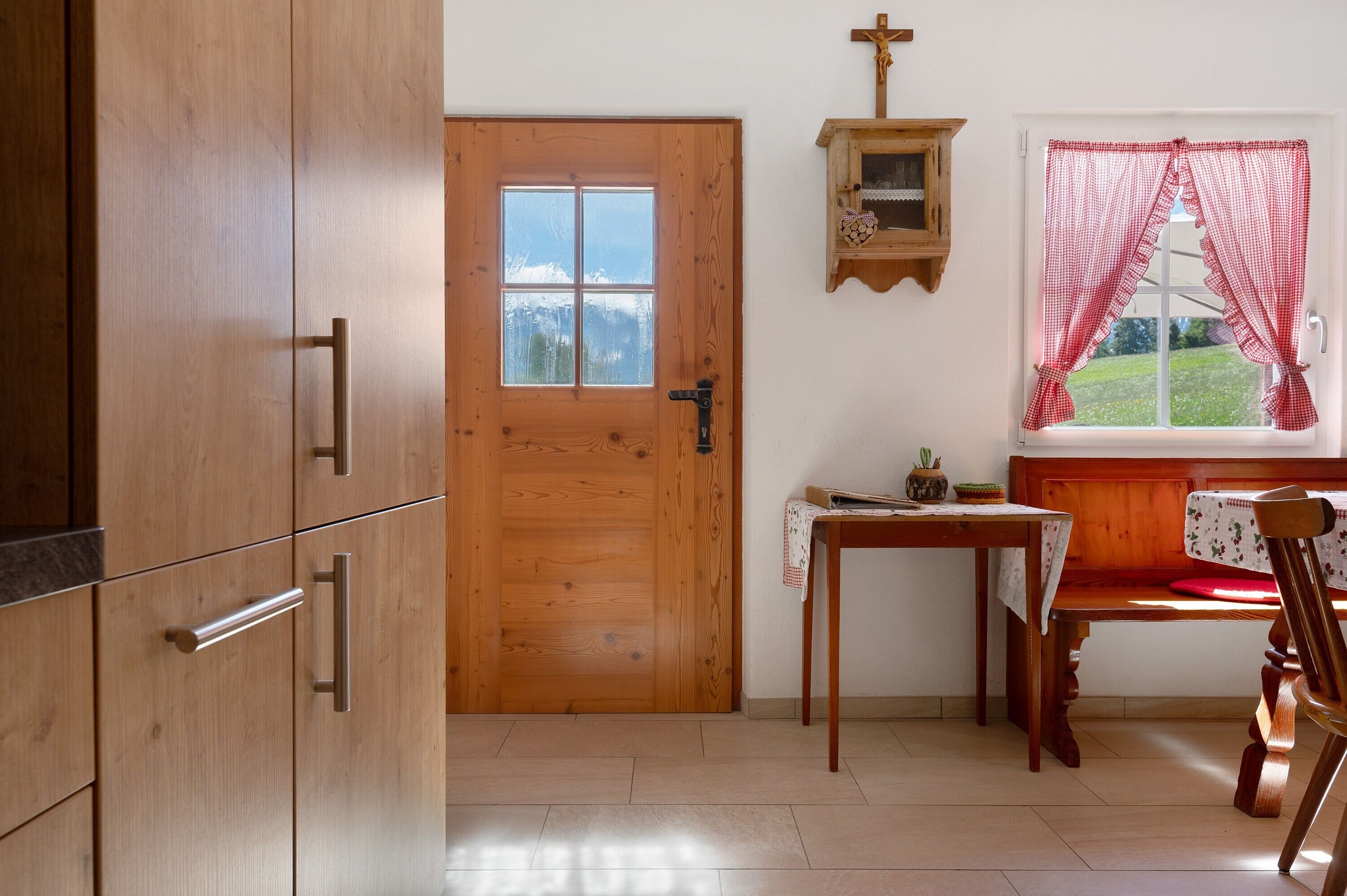 A wooden door, a small table with a chair, a window with red curtains, and a wall-mounted shelf with a cross.