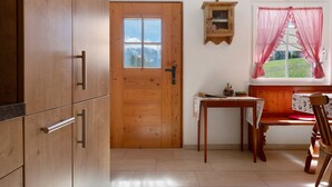 A wooden door, a small table with a chair, a window with red curtains, and a wall-mounted shelf with a cross.