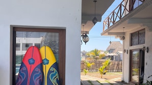 Dos tablas de surf apoyadas contra una pared blanca, una terraza de madera y un camino de piedra que lleva a una casa con balcón.