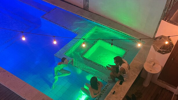 Una piscina con un diseño geométrico de azulejos, rodeada por una terraza de madera y una pequeña área ajardinada.