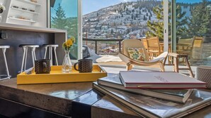 A modern room with a large window overlooking a snowy mountain. There is a yellow tray with a mug and a vase, a chair, and a table with books and a notebook. - Mainstreet Masterpiece Stunning Views! (Park City)