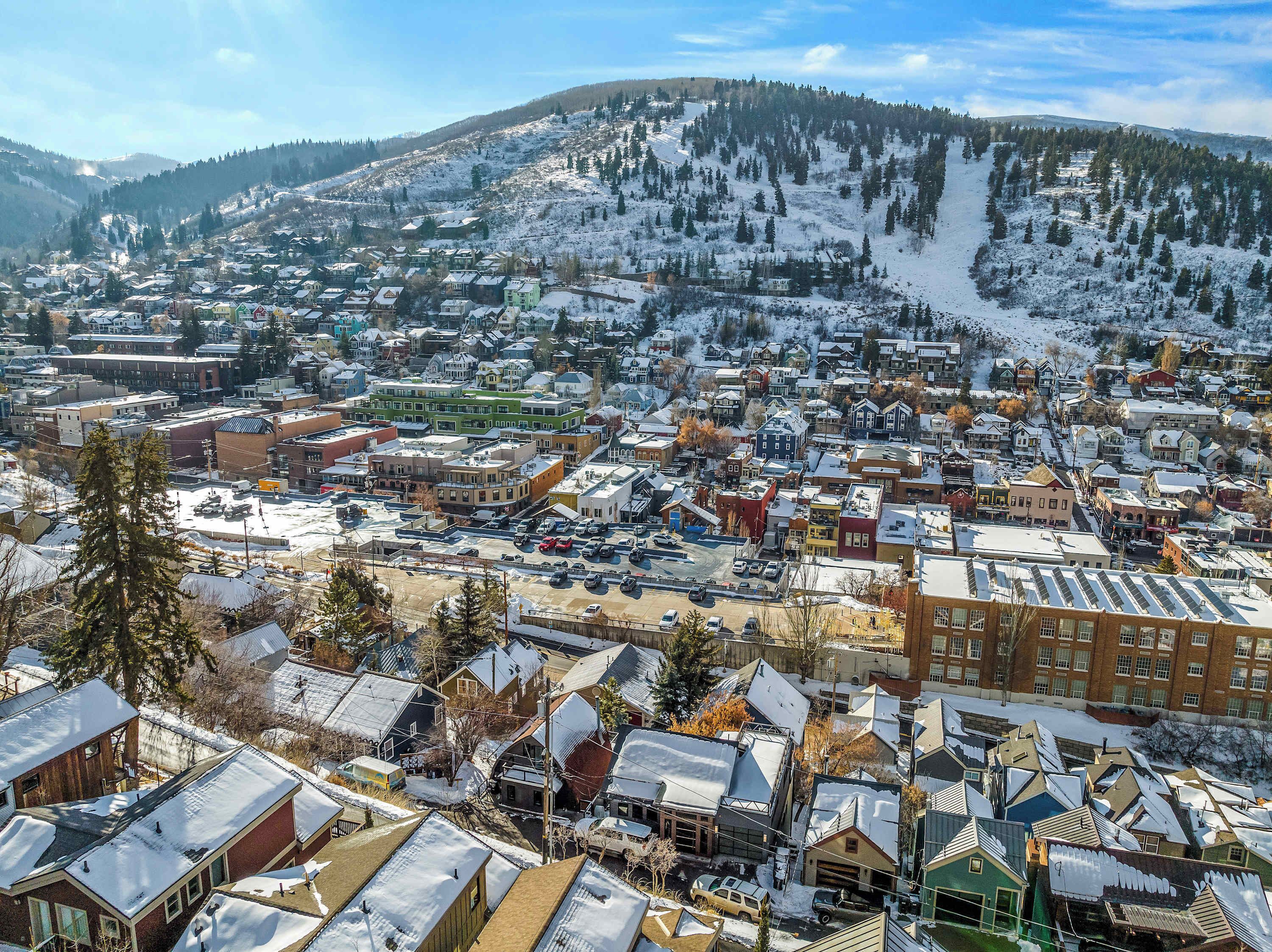 Une ville enneigée, avec un mélange de bâtiments résidentiels et commerciaux, un parking central et, en arrière-plan, des collines recouvertes de neige.